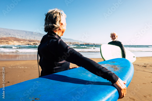 two old and mature people enjoying their vacations outdoors at the beach having fun together with wetsuits and surfboards ready to go surfing - active senior doing water sports activity at summer