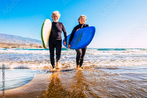 two old and mature people having fun and enjoying their vacations outdoors at the beach wearing wetsuits and holding a surfboard to go surfing in the water with waves - active senior smiling and enjoy