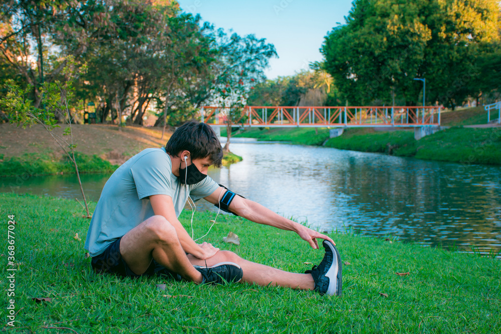 Fototapeta premium A young man doing stretching in a green park before starting to exercise in sportswear and mask.