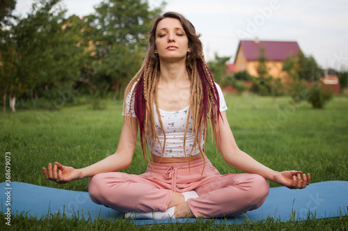 Young beautiful woman with dreadlocks is doing meditation and sitting in lotus position with closed eyes outdoors during morning on green grass of her backyard