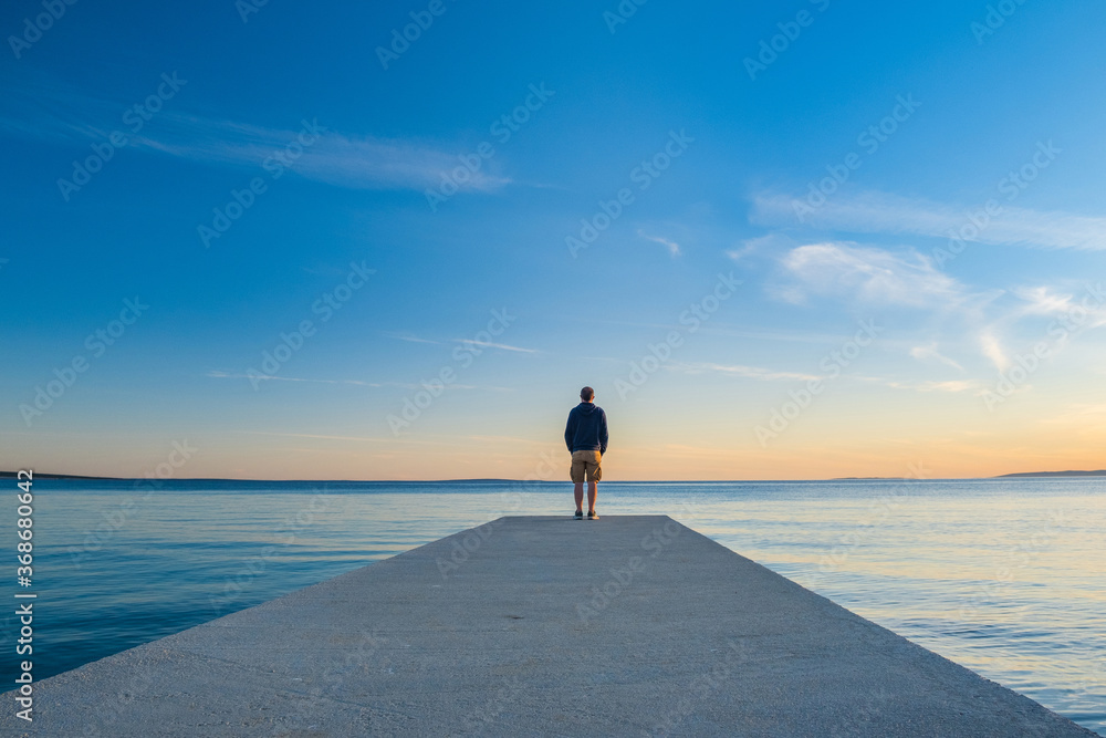 Man in hoodie and short cargo pants standing on dock and looking on sea horizon, island od Pag, Adriatic sea, Croatia