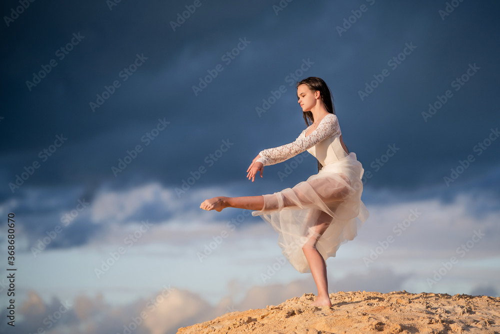 young ballerina in a light long white dress stands in a graceful pose, her skirt and hair develop against the background of the stormy evening sky.