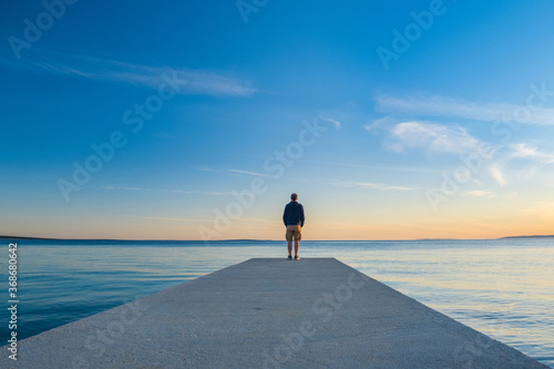 Man in hoodie and short cargo pants standing on dock and looking on sea horizon, island od Pag, Adriatic sea, Croatia 