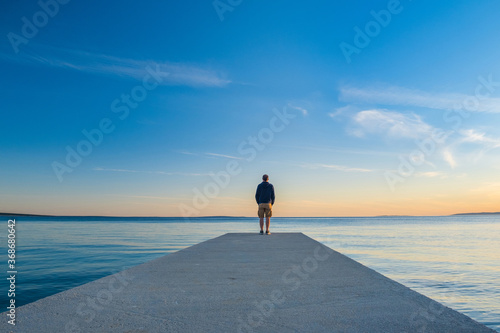 Obraz na płótnie Man in hoodie and short cargo pants standing on dock and looking on sea horizon, island od Pag, Adriatic sea, Croatia
w salonie