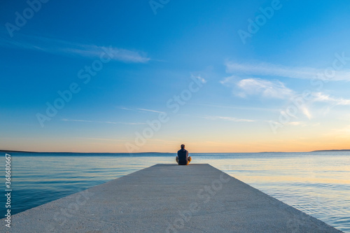 Man in hoodie and short cargo pants sitting on the edge od dock and meditating. Sea horizon, island od Pag, Adriatic sea, Croatia 