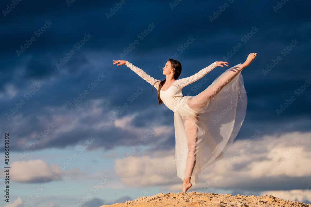 young ballerina in a light long white dress stands in an arabesque like a bird, her skirt develops against the background of the stormy evening sky.