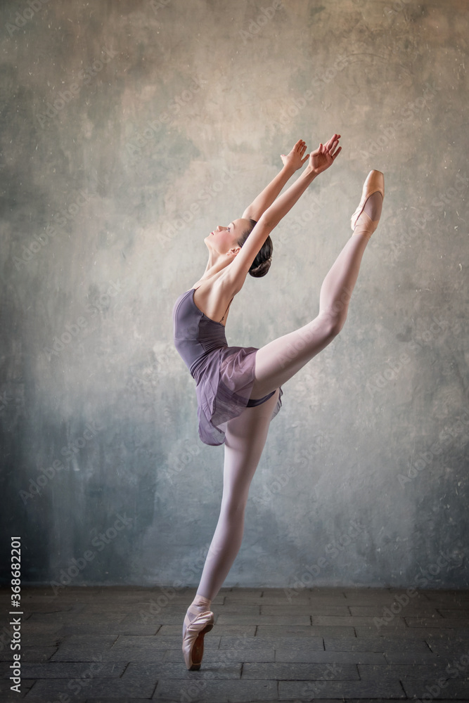 graceful ballet dancer in a light lilac suit and pointe shoes against a gray wall. Dance, grace, artist, contemporary, movement, action and freedom of movement concept.