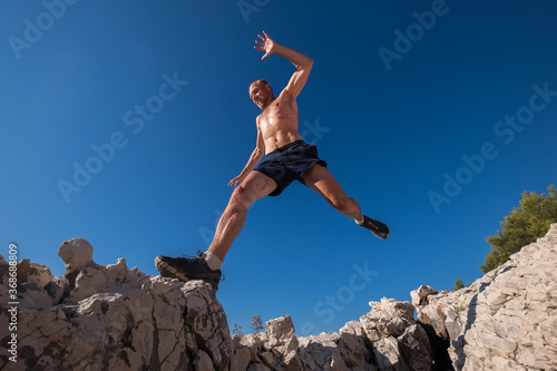 Active fast running mountain sweaty body muscular runner jumping over the cleft cliff during the morning jogging. Sporty people activities wide angle concept image.
