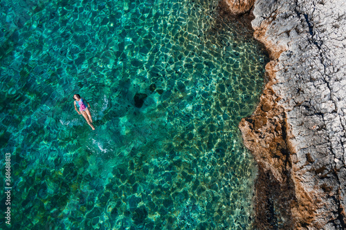 Top aerial view of the young female floating on the back and relaxing on the warm turquoise Adriatic sea waves with rocky coastline. Carefree people vacation time concept image.