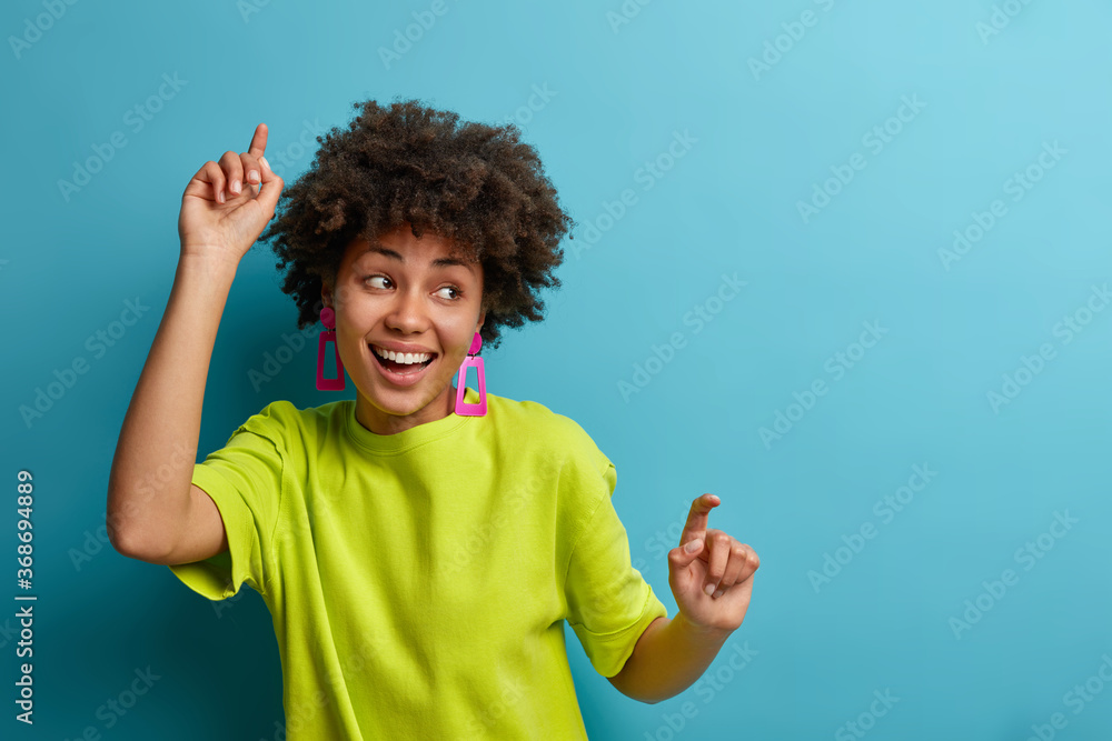 Positive woman with Afro hairstyle dances with arms raised, feels carefree and upbeat, extremly happy and expresses joy, wears green t shirt, isolated over blue background, moves energetically Positive woman with Afro hairstyle dances with arms raised, feels carefree and upbeat, extremly happy and expresses joy, wears green t shirt, isolated over blue background, moves energetically