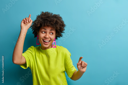 Fotografía Positive woman with Afro hairstyle dances with arms raised, feels carefree and u