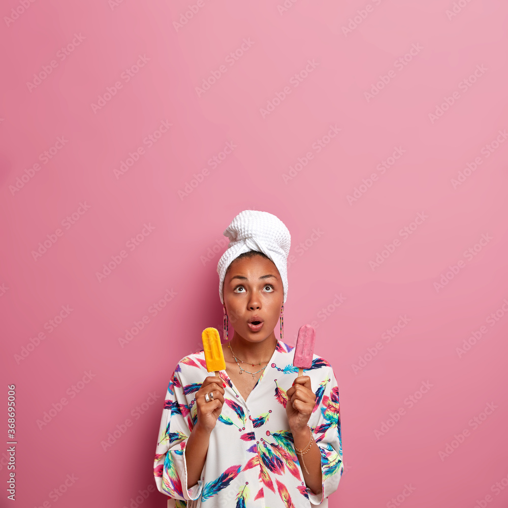 Vertical shot of shocked mixed race woman looking above, holds breath, holds two yummy ice creams of stawberry and mango taste, wears towel and robe after taking bath, has unhealthy nutrition Vertical shot of shocked mixed race woman looking above, holds breath, holds two yummy ice creams of stawberry and mango taste, wears towel and robe after taking bath, has unhealthy nutrition