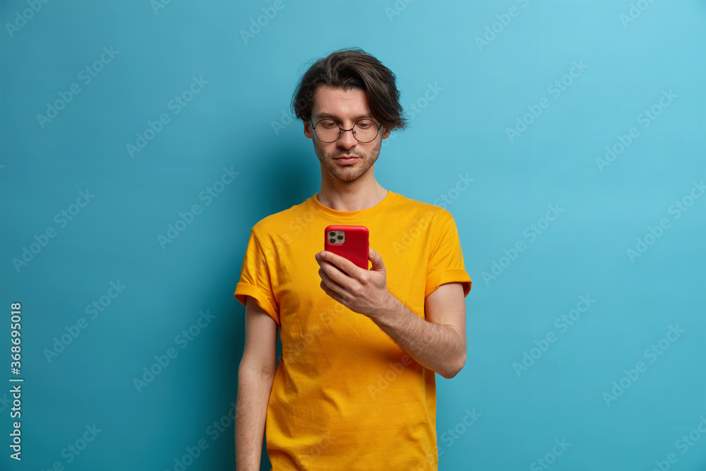 Serious unshaven man holds cell phone in hand, concentrated in screen, watches tutorial video, types answer or reads feedback about new project, wears transparent glasses, isolated on blue wall Serious unshaven man holds cell phone in hand, concentrated in screen, watches tutorial video, types answer or reads feedback about new project, wears transparent glasses, isolated on blue wall
