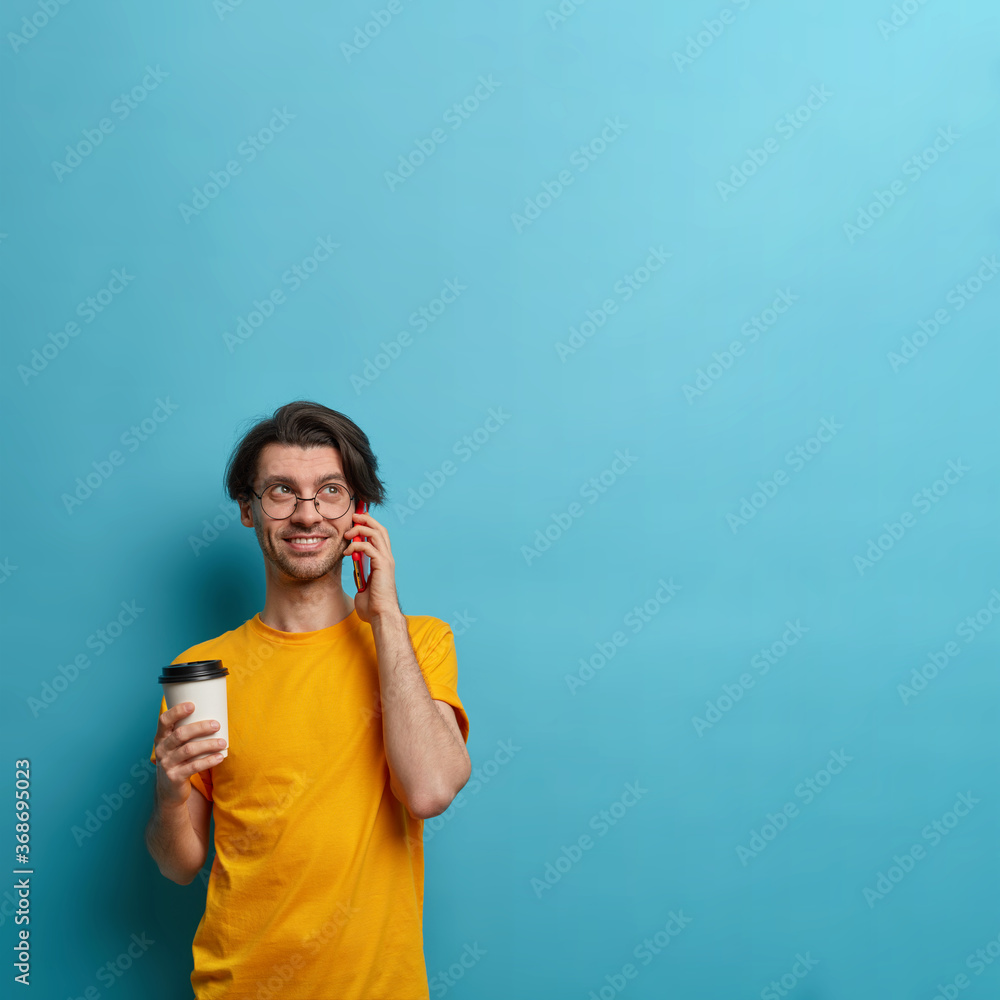 Smiling guy keeps cellular near ear, busy talking with friend, discusses good news, holds takeaway cup of coffee, communicates pleasantly, poses against blue backgroud, copy space for your information Smiling guy keeps cellular near ear, busy talking with friend, discusses good news, holds takeaway cup of coffee, communicates pleasantly, poses against blue backgroud, copy space for your information