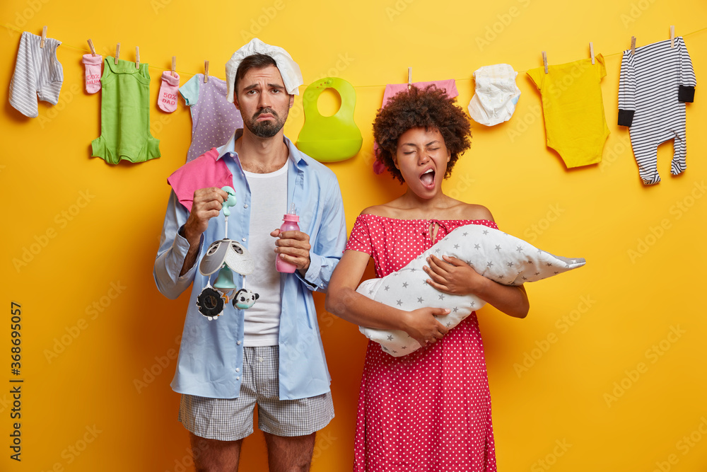 Young tired exhausted parents spend sleepless night caring about infant, nursing baby. Sleepy mother yawns and holds child on hands wrapped in blanket, displeased father holds bottle and mobile toy