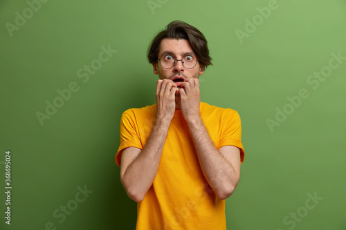 Photo of worried nervous man bites finger nails and stares with scared expression, frightened by something terrifying, wears round spectacles and yellow t shirt, poses against green background