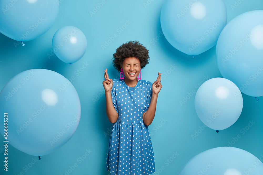 Fototapeta premium Happy celebration concept. Positive hopeful birthday girl crosses fingers, makes wish, believes all her dreams come true dressed in polka dot dress in one tone of background. Inflated balloons around