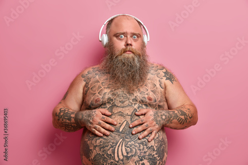 Studio shot of surprised guy keeps hands on tummy, has tattooed stomach and arms, stands with naked body, being overweight, listens favouite audio track in headphones, isolated on pink wall.