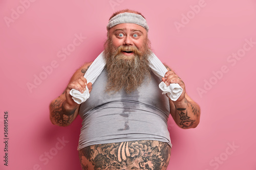 Portrait of fat bearded man with sweaty body, feels tired after exhausting exercises in gym, has big belly sticking out from t shirt, keeps hands on towel, does sport regularly to loose weight