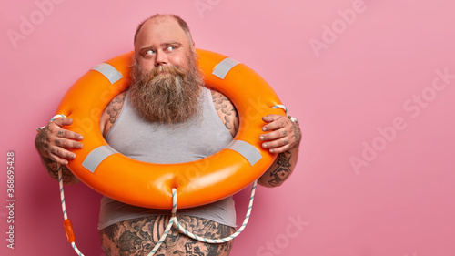 Summer rest concept. Pensive European man with thick beard, dressed in undersized shirt, has big tummy, poses with lifebuoy, wants to swim, isolated on pink background, empty space for text.