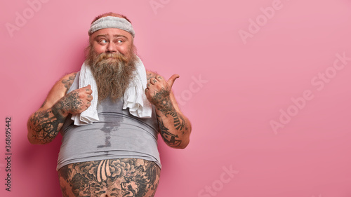 Purposeful persistent motivated stout guy has regular workout at gym, wears hairband, undersized vest and towel around neck, has big abdomen with tattoo, points thumb on blank space for your text