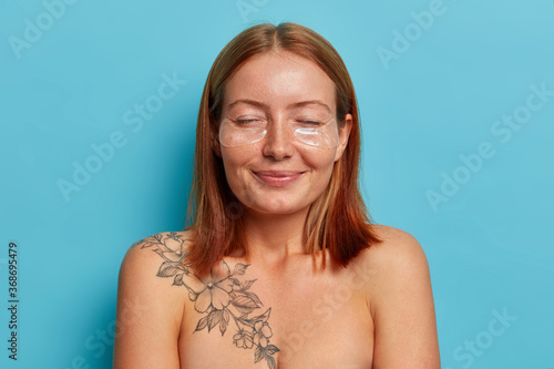 Women, face care and beauty concept. Cheerful smiling redhead woman stands with eyes closed, applies hydrogel patches, has smooth clean skin, well cared body, poses naked against blue background