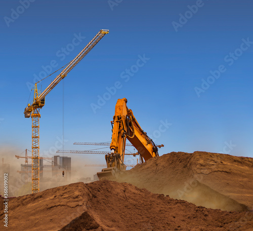 A dusty construction site with a yellow crane and an excavator bucket under a clear blue summer sky  creating copy space for text