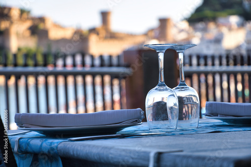 Fototapeta Naklejka Na Ścianę i Meble -  Table covered by a blue tablecloth, with two glasses and two plates with knives and forks. Restaurant in front of the sea, with a blurred background of the medieval castle of the town Tossa de Mar.