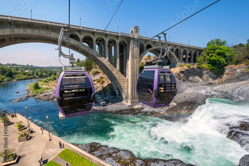 Fototapeta Naklejka Na Ścianę i Meble -  Two gondolas fly over the Spokane River and falls near Riverfront Park on a summer day in Spokane, Washington, USA