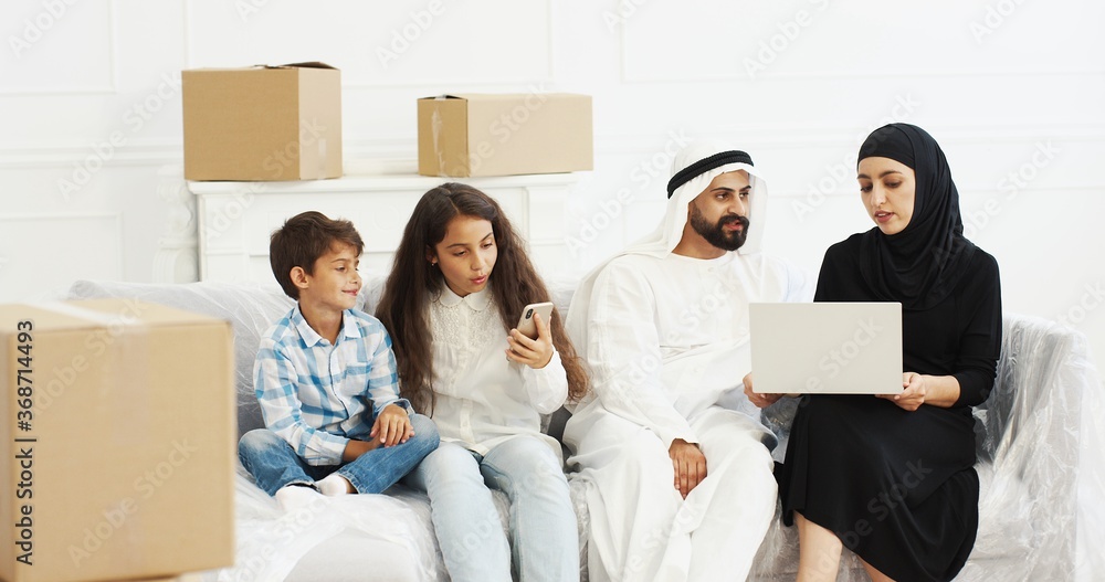 Arabian young parents in dishdasha and hijab sitting on couch with laptop computer and planning interior for new home after moving in. Small kids among boxes playing on smartphone. Arabs with children Arabian young parents in dishdasha and hijab sitting on couch with laptop computer and planning interior for new home after moving in. Small kids among boxes playing on smartphone. Arabs with children