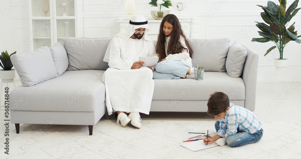 Arabian father in white kandura playing with small kids, daughter and son in living room. Muslim dad drawing and coloring pictures. Boy and girl with daddy painting at home and spending time together. Arabian father in white kandura playing with small kids, daughter and son in living room. Muslim dad drawing and coloring pictures. Boy and girl with daddy painting at home and spending time together.