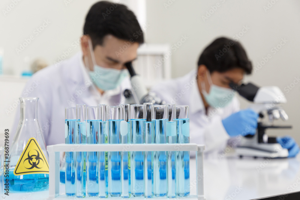 Focus on Solution Tubes in front of Two Male Scientists wear Face Mask working in Lab while Checking Solution by micro scope On Background. Focus on Solution Tubes in front of Two Male Scientists wear Face Mask working in Lab while Checking Solution by micro scope On Background.