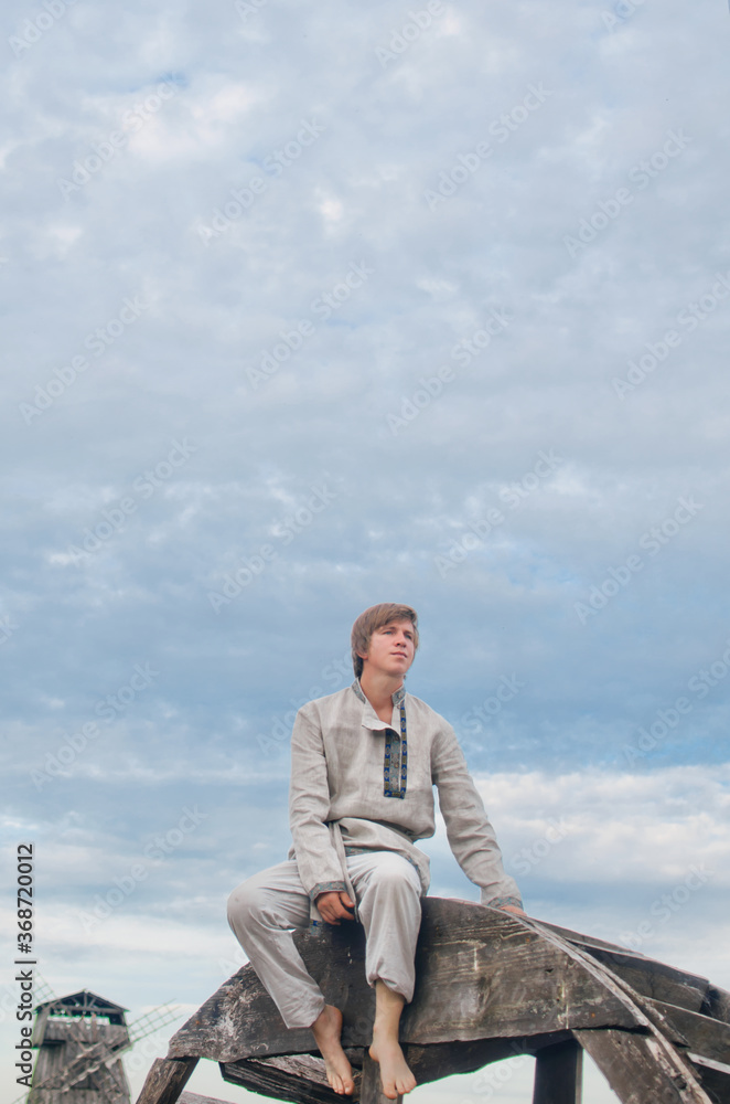 a young man in a national costume of white linen sits on an old water mill wheel and looks into the distance a young man in a national costume of white linen sits on an old water mill wheel and looks into the distance