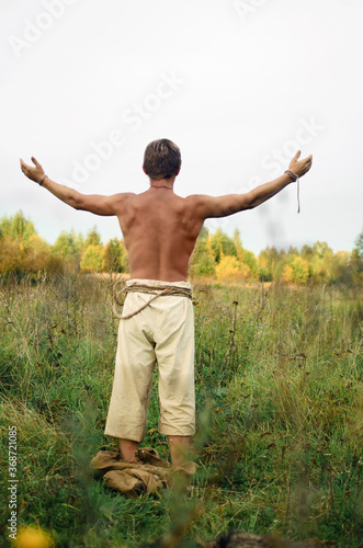 young man stripped to the waist stands in a wild field with his arms outstretched