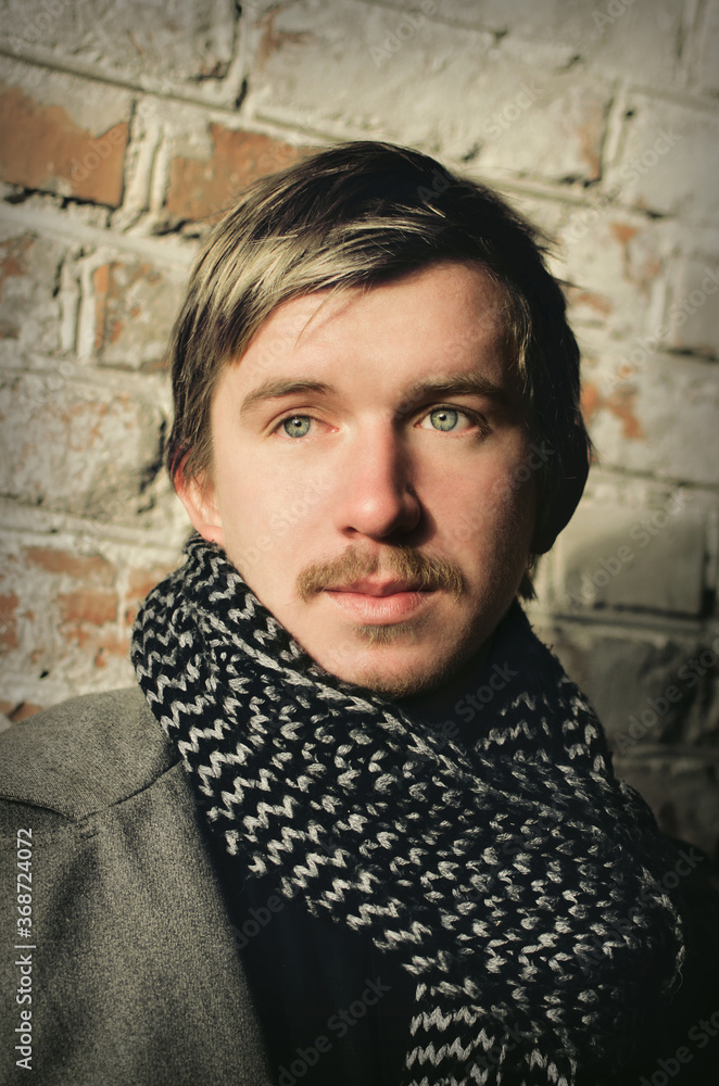 portrait of a young mustachioed guy in a warm coat and scarf against an old brick wall