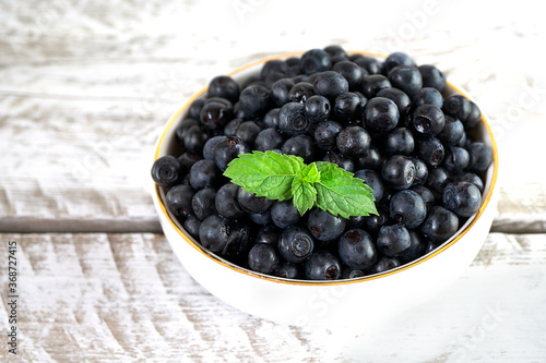 Delicious and healthy blueberries in a white bowl stand on a vintage white wooden table. The bowl is decorated with fresh mint leaves. Seasonal summer berries, healthy vitamin dessert. Top view.