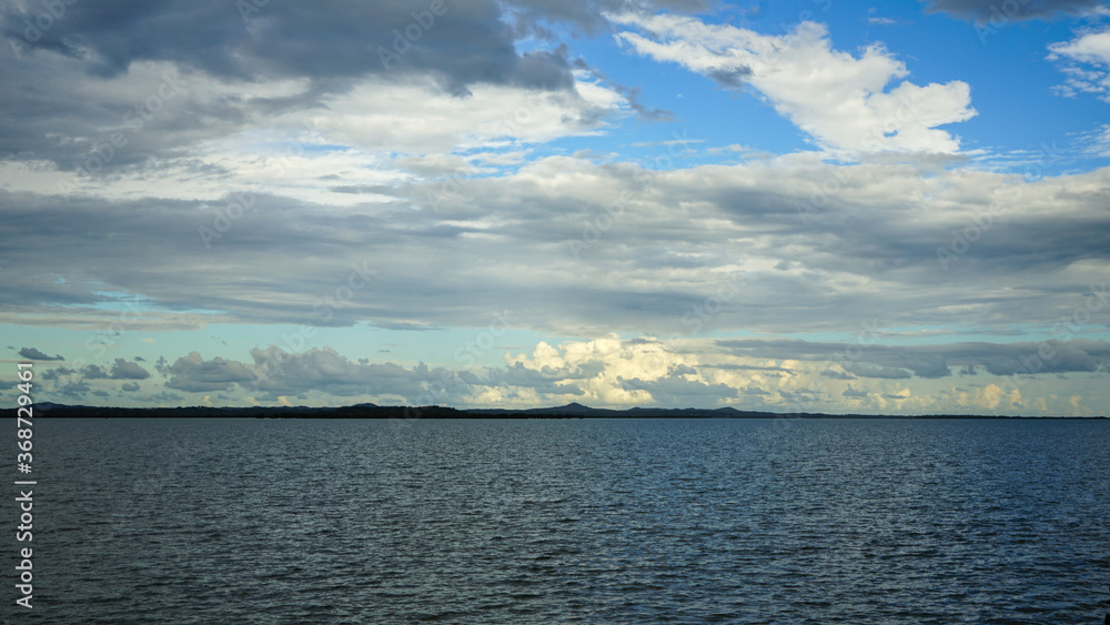 Cloudscape with grey and white clouds against a blue sky fading to pale aqua near the horizon, with blue-grey sea in the foreground and land on the horizon. Moreton Bay, Queensland, Australia.