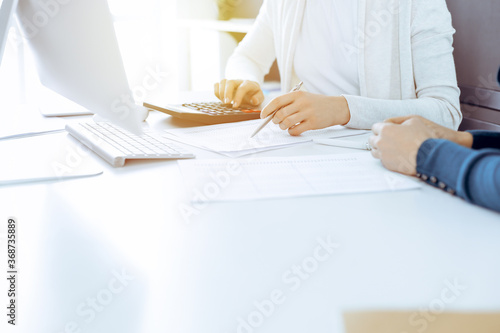 Accountant checking financial statement or counting by calculator income for tax form, hands close-up. Business woman sitting and working with colleague at the desk in office. Audit concept