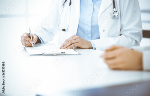 Unknown woman-doctor and female patient sitting and talking at medical examination in clinic  close-up. Therapist wearing blue blouse is filling up medication history record. Medicine concept