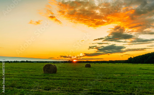 Scenic view at picturesque sunset in a green shiny field with hay stacks, bright cloudy sky , trees and golden sun rays with glow, summer valley landscape