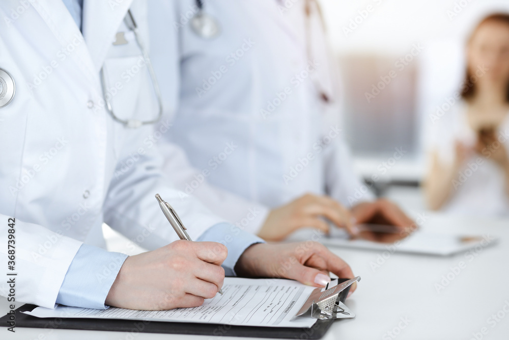 Unknown woman-doctors at work with patient at the background. Female physicians filling up medical documents or prescription while standing in sunny hospital reception desk, close-up. Health care Unknown woman-doctors at work with patient at the background. Female physicians filling up medical documents or prescription while standing in sunny hospital reception desk, close-up. Health care
