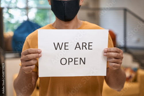 Business after lockdown. Close up shot of a male office worker in black protective mask showing paper with text WE ARE OPEN at camera, standing in the office