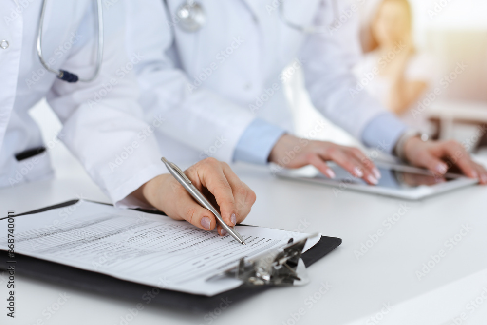 Unknown woman-doctors at work with patient at the background. Female physicians filling up medical documents or prescription while standing in hospital reception desk, close-up. Health care concept Unknown woman-doctors at work with patient at the background. Female physicians filling up medical documents or prescription while standing in hospital reception desk, close-up. Health care concept
