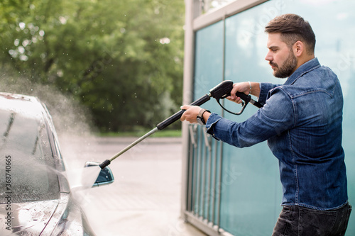 Handsome bearded young man in jeans shirt, washing his new car manually with water high-pressure hose at outdoor self wash service. Car washing concept.