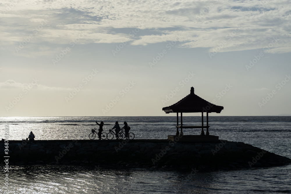 Silhouette of cyclists taking selfie near the sea. Cycling together with friends at the beach with flat sky at stone deck and balinese building