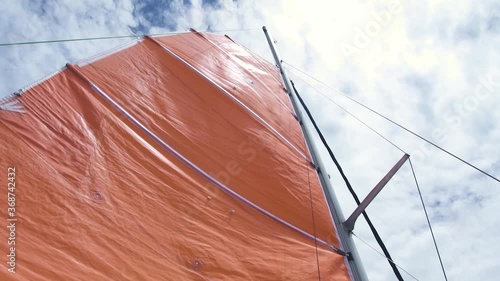 Mast and mainsail of a sailboat with ropes against the white clouds, view from below. Orange mainsail and metal mast. Shot from lower anlge on sunny day. photo