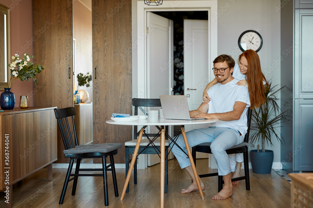 adorable female hugs her working husband from back, they look at screen of laptop and smile, at home in domestic wear adorable female hugs her working husband from back, they look at screen of laptop and smile, at home in domestic wear