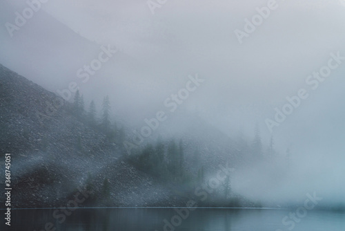 Ghostly atmospheric view to tranquil mountain lake and stony steep slope with coniferous trees in dense fog. Minimalist scenery with low clouds and calm water. Mysterious place at early foggy morning.