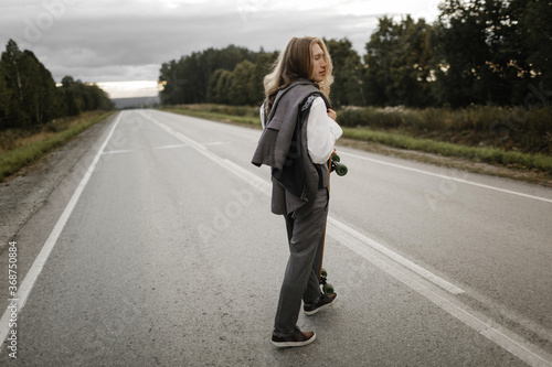 Man with long blond hairs in grey office suit with skateboard longboard in hands is walking down road in city outskirts. He is going to ride a skateboard  front view. Freedom from office work concept.