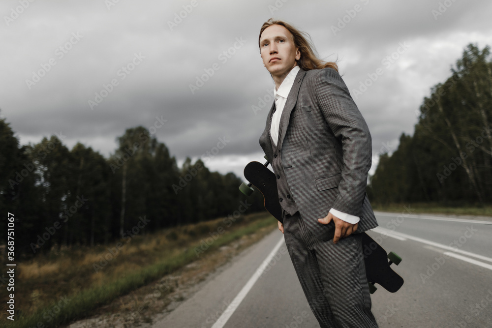 Man with long blond hairs in grey office suit with skateboard longboard in hands is walking down road in city outskirts. He is going to ride a skateboard, front view. Freedom from office work concept. Man with long blond hairs in grey office suit with skateboard longboard in hands is walking down road in city outskirts. He is going to ride a skateboard, front view. Freedom from office work concept.